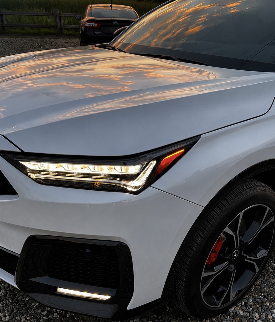Close-up of a white Acura MDX hood and headlight reflecting sunset clouds after coating